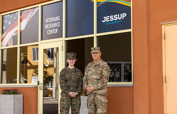 2 Veterans (female and male) stand in front building in full uniform smiling.