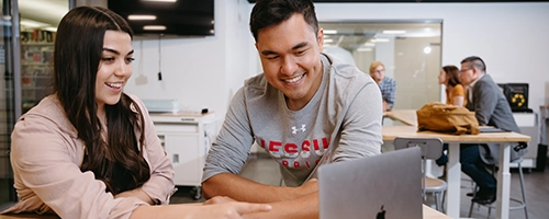 Two students (male and female) looking at computer together and smiling.